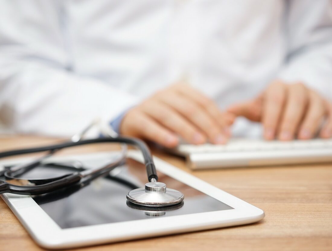 Blurred doctor in background typing on computer keyboard with tablet and stethoscope in foreground
